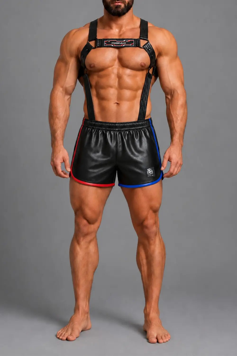 A buff sports physique man modeling black leather-like athletic shorts with red and blue side trim, plus a black harness-style top with shoulder straps, against a plain gray studio background.